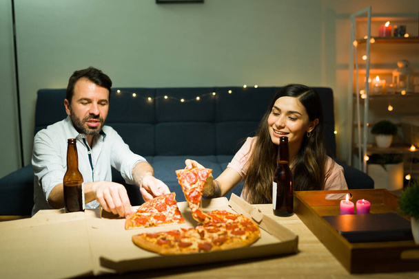 Hungrig fühlen. Schöne Freundin und schöner Freund, der Pizza isst und Bier trinkt. Latino-Paar entspannt sich zu Hause bei einem Date am Abend  - Foto, Bild