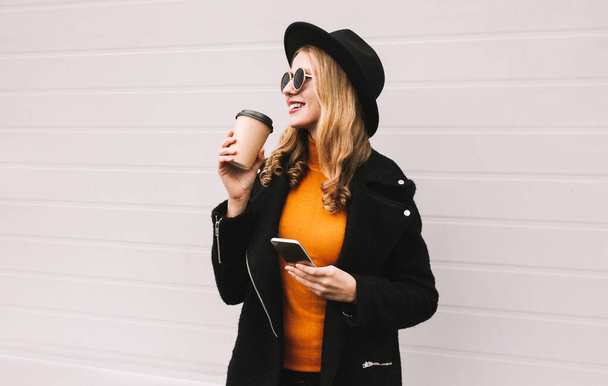 Elegante ritratto di bella giovane donna che beve caffè con smartphone guardando altrove indossando un cappotto nero e cappello rotondo su sfondo grigio - Foto, immagini