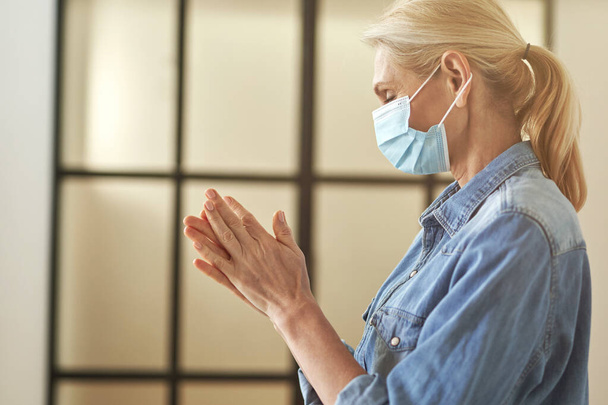 Side view of mature blonde woman in protective face mask rubbing, cleaning her hands using antibacterial hand sanitizer, standing indoors - Foto, immagini
