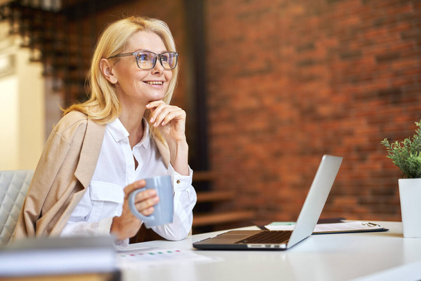 Fröhliche reife Geschäftsfrau mit Brille, die weglächelt, Laptop benutzt und während der Arbeit im Büro Kaffee trinkt - Foto, Bild