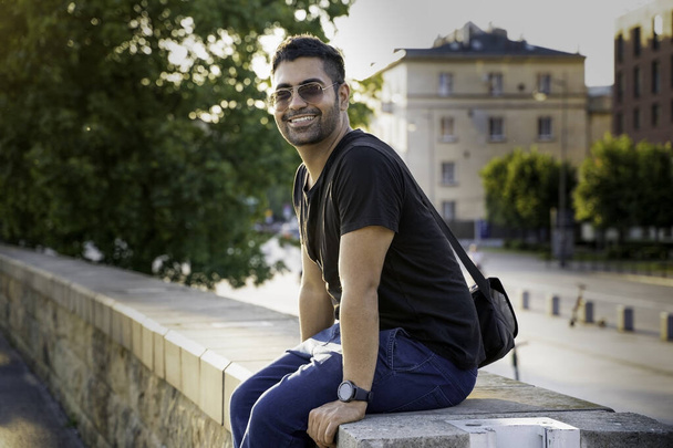 Indian arab or hispanic young man in black shirt with a shoulder strap bag and sunglasses sitting on a wall smiling while looking at camera against blurred background with European architecture - Photo, Image