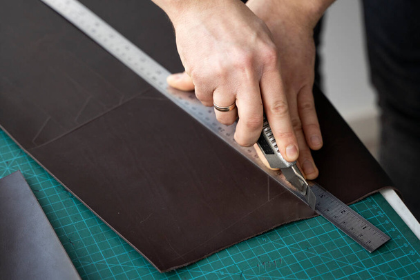 Mens hand holding a stationery knife and metal ruler and cutting on a pieces for a leather wallet in his workshop. Working process with a brown natural leather. Craftsman holding a crafting tools. - Foto, immagini