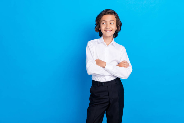 Foto de divertido adorable niño de la escuela usar traje formal sonriendo brazos plegados buscando espacio vacío aislado color azul fondo - Foto, Imagen