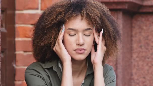 Face of unhappy young African American woman siting outdoors rubs temple, closed eyes suffers headache painful feelings chronic migraine hurt. Break up, difficult period of life concept closeup view - Footage, Video