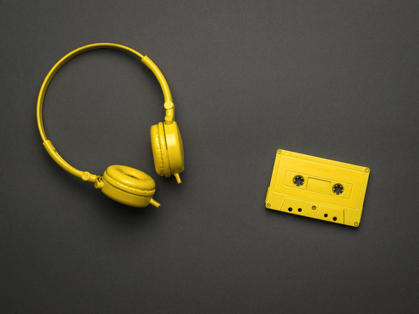 Auriculares amarillos clásicos y un casete amarillo con una cinta magnética sobre un fondo negro. Tendencia de color. Equipo vintage para escuchar música. Puesta plana. - Foto, Imagen