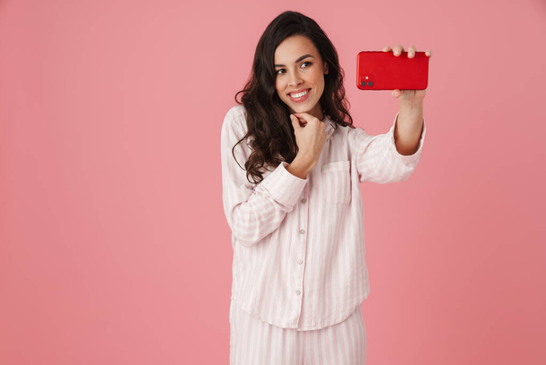 Cheerful beautiful woman taking selfie on mobile phone isolated over pink background - Fotografie, Obrázek