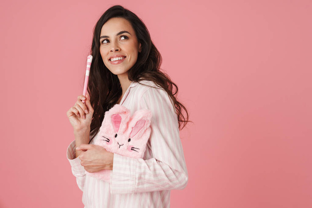 Happy beautiful woman in pajama posing with pen and bunny diary isolated over pink background - Photo, image