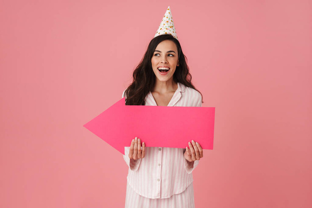 Joyful woman in party cone laughing while posing with arrow placard isolated over pink background - Fotoğraf, Görsel