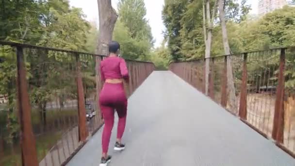 Mujer de fitness negro en ropa deportiva corriendo en el puente al aire libre durante el entrenamiento diario por la mañana. Joven atlética hermosa hembra cuida de su cuerpo sano, quiere perder peso, trotar. Vista trasera - Metraje, vídeo