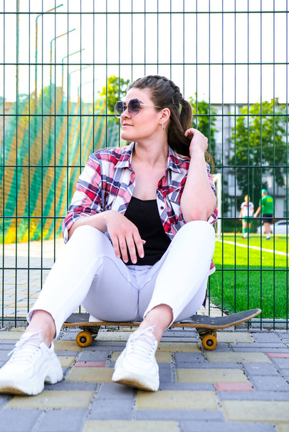 Young millennial woman hippie sitting on skateboard. Summer Skate Sessions. Woman cruising on a longboard in a park. Carefree female skater 20s enjoying freedom youth lifestyle - 写真・画像