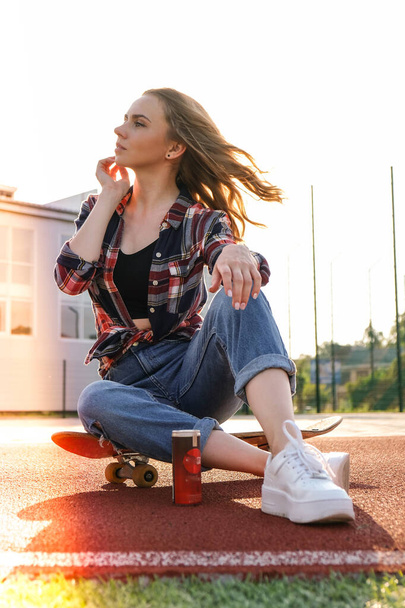 Mladá tisíciletá hippie sedí na skateboardu. Letní brusle. Žena křižující na dlouhém prkně v parku. Carefree žena bruslařka 20s těší svobodu mládež životní styl - Fotografie, Obrázek