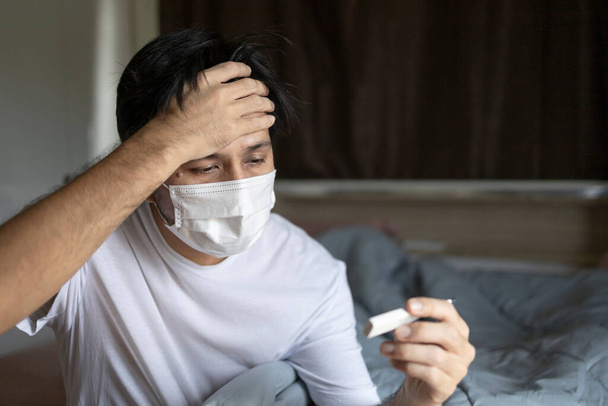 Asian sick man wearing face protection mask and holding thermometer having fever in the bedroom at home - Photo, Image