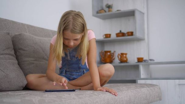 Ένα κορίτσι σχολικής ηλικίας είναι στο σπίτι και χρησιμοποιεί έναν υπολογιστή tablet για να παίξει και να ζωγραφίσει. Απομακρυσμένη εκπαίδευση χωρίς να φύγεις. Ανάπτυξη ικανοτήτων στα παιδιά. - Φωτογραφία, εικόνα