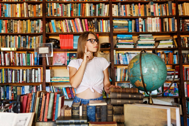 Молодой улыбающийся привлекательный портрет студентки с очками, стоящими в библиотеке, с книгами в руках.. - Фото, изображение