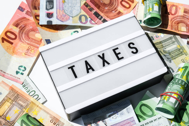Доска с надписью TAXES черными буквами вокруг банкнот евро. Концепция налоговых платежей и подачи документов. Деньги, бизнес, финансы, инвестиции, сбережения и коррупция. Денежный счет - Фото, изображение