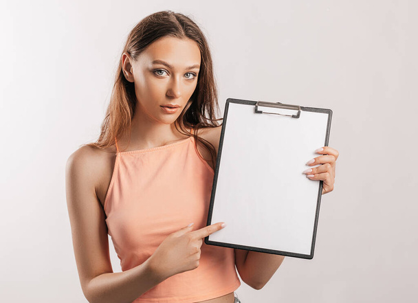 Girl student points to the sheet. Beautiful young business woman holds a clipboard with mockup blank space isolated on gray background. Study and business concept. Online learning cover - Foto, afbeelding