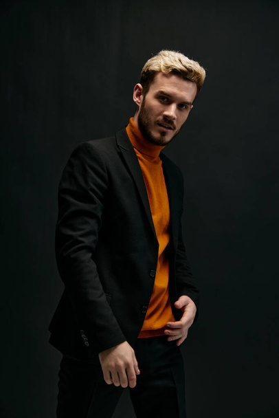 Stylisches Männermodel in orangefarbenem Pullover und Jacke hält seine Hand am Gürtel vor dunklem Hintergrund. Hochwertiges Foto - Foto, Bild