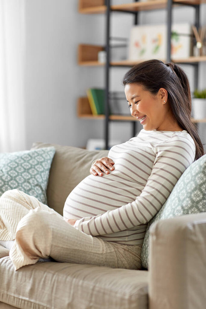 happy pregnant asian woman sitting on sofa at home - Foto, Imagem