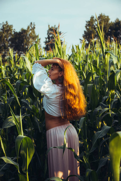 Young beautiful woman with long red hair between green leaves on a corn field in summer, in the light of the setting sun. - Φωτογραφία, εικόνα