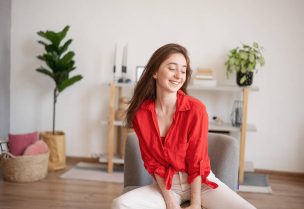 Portrait of a young happy brunette woman in a red shirt sitting on a chair at home in the interior - Photo, image