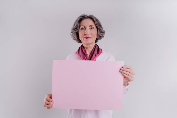 Mulher sênior elegante feliz segurando rosa cartão de papel vazio no fundo cinza claro com espaço em branco para o texto. - Foto, Imagem