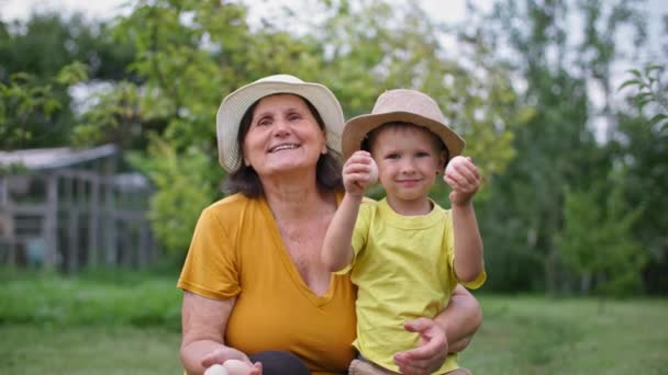 huevos de granja caseros orgánicos, abuela con su nieto en sombreros de paja posando para la cámara con huevos en las manos en un jardín verde - Imágenes, Vídeo