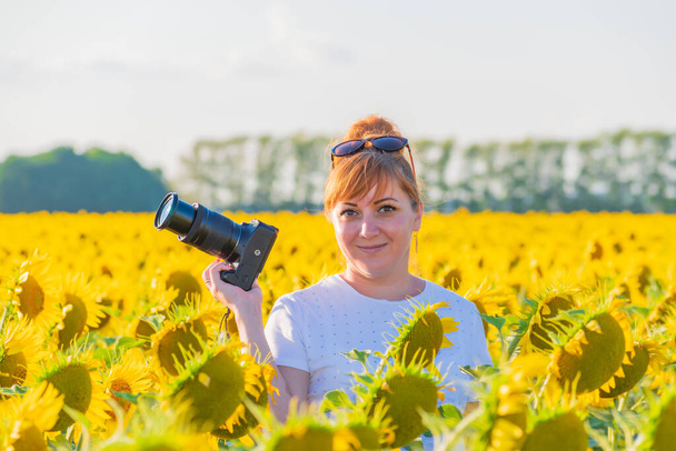 Elinde kamera olan genç kızıl saçlı kadın fotoğrafçı sarı bir ayçiçeği tarlasında duruyor. Kameraya bak.. - Fotoğraf, Görsel