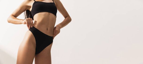 Cropped studio image of female body with slim figure in black underwear posing over white studio background. Flyer - Foto, Bild