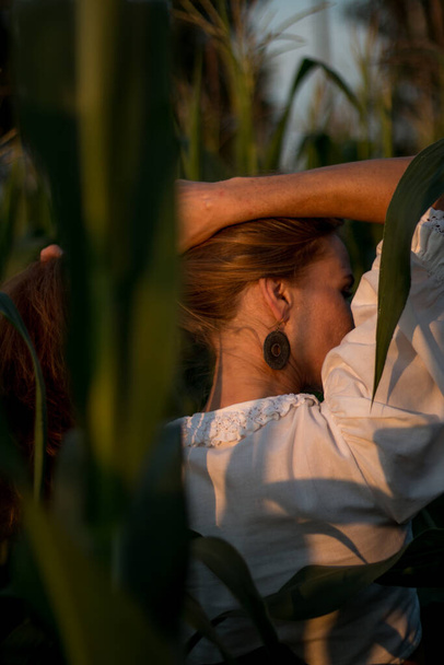 Young beautiful woman with long red hair between green leaves on a corn field in summer, in the light of the setting sun. - Photo, image