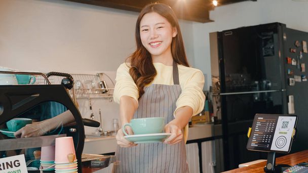 Портрет молодой азиатской официантки-баристы, держащей чашку кофе, чувствующей себя счастливой в городском кафе. Девушка в фартуке с зубастой улыбкой смотрит на стойку фотоаппарата в кофейне. - Фото, изображение