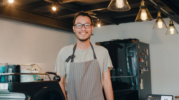 Portrét mladý Asiat barista pocit, šťastný úsměv v městské kavárně. Malý podnikatel majitel japonský muž v zástěře uvolnit zubatý úsměv při pohledu do kamery stojící u pultu v kavárně. - Fotografie, Obrázek