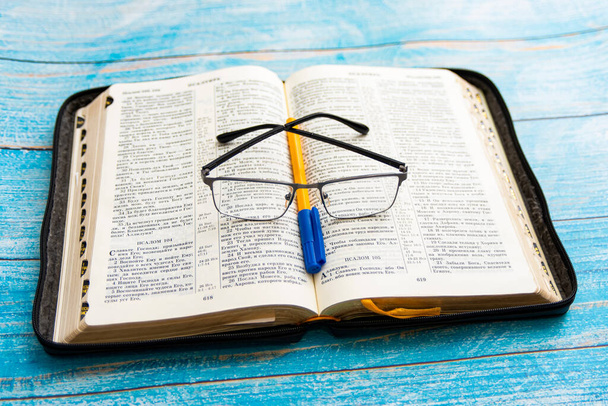 Στην ανοιχτή Βίβλο βρίσκεται ένα στυλό με γυαλιά, ένα μπλε φόντο. - Φωτογραφία, εικόνα