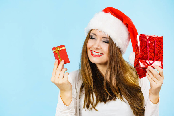 Χριστουγεννιάτικα δώρα. Ευτυχισμένη γυναίκα φορώντας καπέλο santa claus κρατώντας την παρούσα κόκκινη τσάντα και μικρό κουτί δώρου με κοσμήματα, σε μπλε - Φωτογραφία, εικόνα