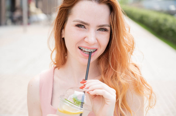 Young beautiful red-haired woman with braces drinks cooling cocktail outdoors in summer. Portrait of a smiling girl with freckles - Foto, imagen
