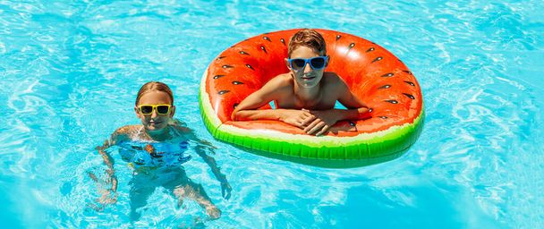 Happy kids, boy and girl in sunglasses, swim and play with inflatable rings, have fun and splash water in a beautiful pool in a tropical resort, have fun during family summer vacation - Photo, image