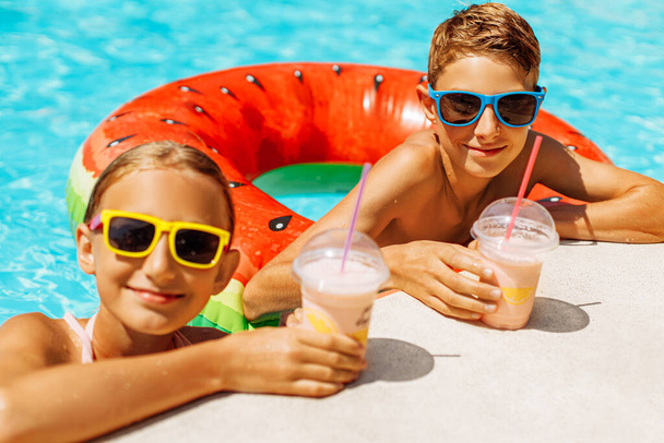 Close-up of cheerful little boy and girl in sunglasses drinking cocktails while enjoying in the pool, Cute brother and sister leaning on the edge of the pool and smiling, Summer holiday concept - Photo, image