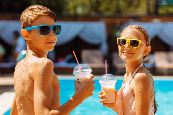 Happy cute kids, boy and girl drinking cool summer cocktails, having fun and relaxing sitting near the pool with clear blue water, happy summer, friendly family weekend concept, outdoor lifestyle - Photo, image