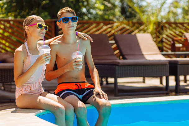 Happy cute kids, boy and girl drinking cool summer cocktails, having fun and relaxing sitting near the pool with clear blue water, happy summer, friendly family weekend concept, outdoor lifestyle - Foto, Imagem