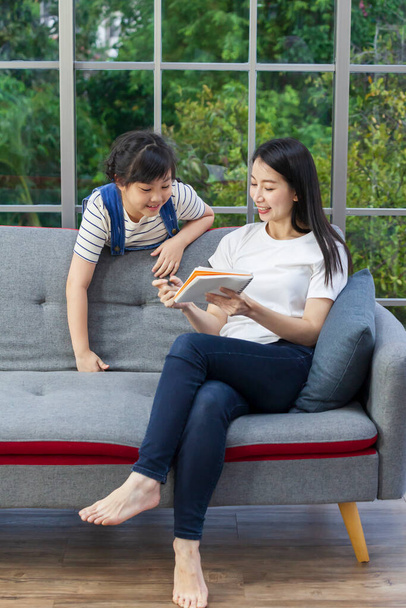 Asian mother teaching homework to daughter while sitting on the sofa couch at home for homeschooling and education concept - Photo, Image