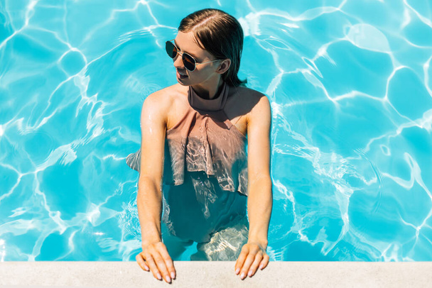 Beautiful young tanned woman in sunglasses, in the outdoor pool, woman in a swimsuit sunbathing in the pool, Happy woman relaxing in the pool, The concept of summer, vacation, travel - Photo, Image