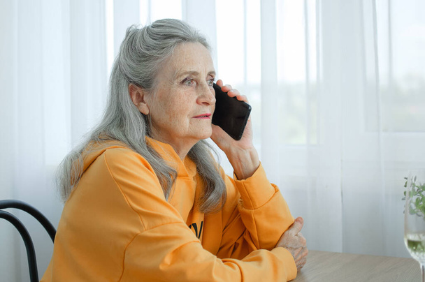 Beautiful old grandmother with grey hair and face with wrinkles is using smartphone, talking with someone and sitting at the table at home on window background, mothers day, happy retirement - Photo, image