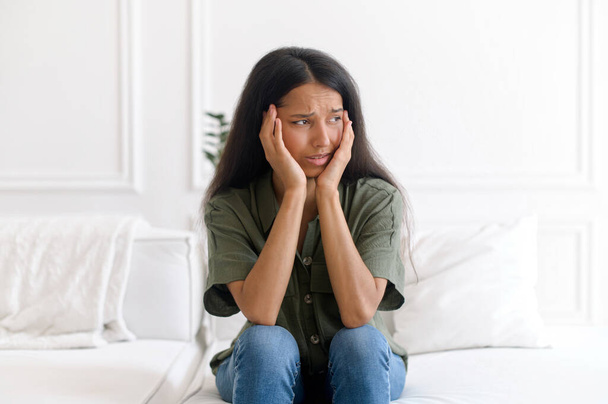 Scared indian woman feels panic attack, sits on the sofa at home - Fotó, kép