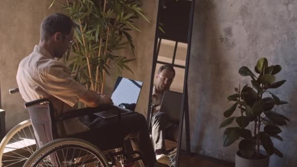 Πλήρης πλαϊνή άποψη των ατόμων με ειδικές ανάγκες Καυκάσιος άνδρας σε αναπηρική καρέκλα, χρησιμοποιώντας φορητό υπολογιστή μπροστά από καθρέφτη στο δωμάτιό του - Πλάνα, βίντεο