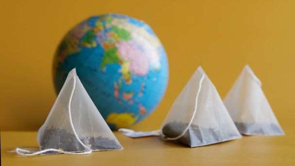  果実や果実の入った紅茶が入った白いピラミッド型のティーバッグは、黄色の背景に地球の隣にあります。世界中のお茶の人気の概念。選択的集中。閉鎖 - 写真・画像