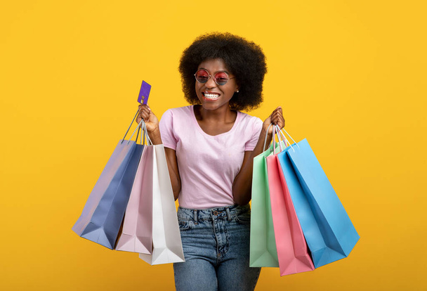 Cliente feliz. Joyful senhora preta com sacos de compras e cartão de crédito ilimitado sorrindo sobre fundo amarelo - Foto, Imagem