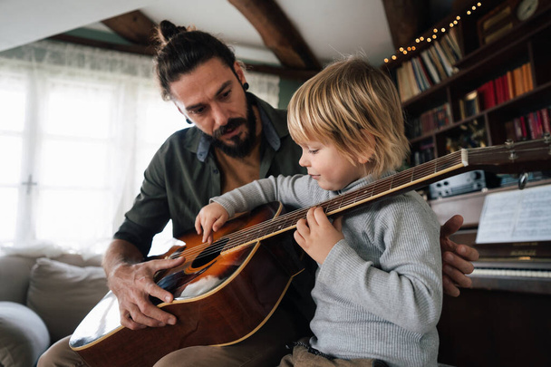 Cute little boy and his handsome father are playing guitar and smiling at home. Family love concept - Fotografie, Obrázek