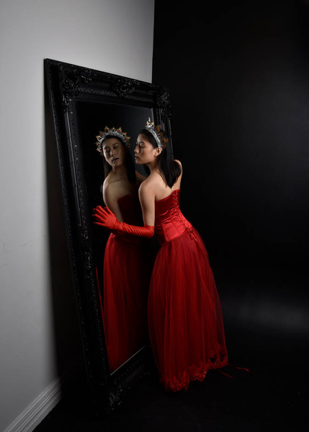Ritratto a figura intera di bella giovane donna asiatica che indossa corsetto rosso, lunghi guanti da opera e copricapo decorato corona. Graceful posa contro uno specchio a figura intera con uno sfondo scuro studio. - Foto, immagini