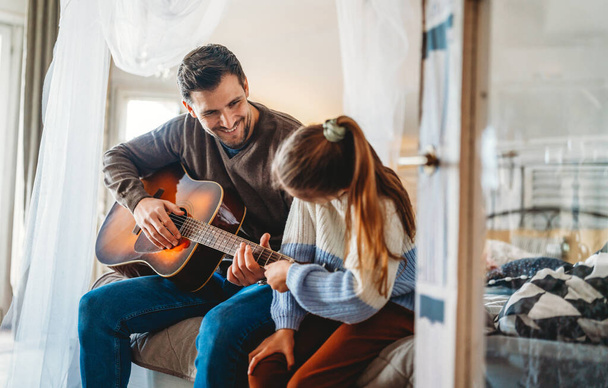 Happy girl and her handsome father are playing guitar and smiling while sitting on couch at home - Фото, зображення