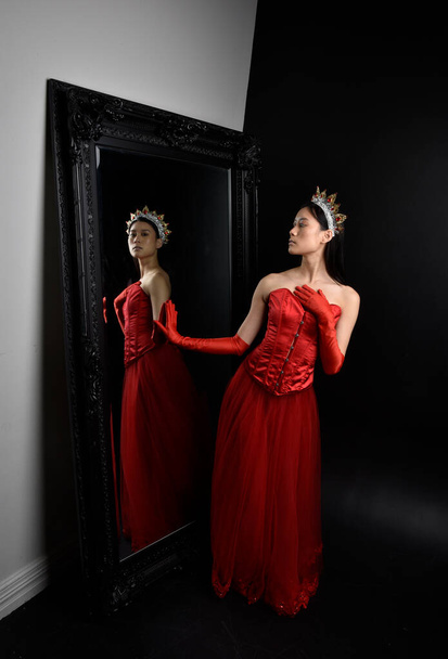 Πλήρες πορτραίτο μιας όμορφης νεαρής Ασιάτισσας με κόκκινο κορσέ, μακριά γάντια όπερας και περίτεχνη κόμμωση στέμματος. Χαριτωμένη ποζάροντας σε έναν ολόσωμο καθρέφτη με σκοτεινό φόντο στούντιο. - Φωτογραφία, εικόνα