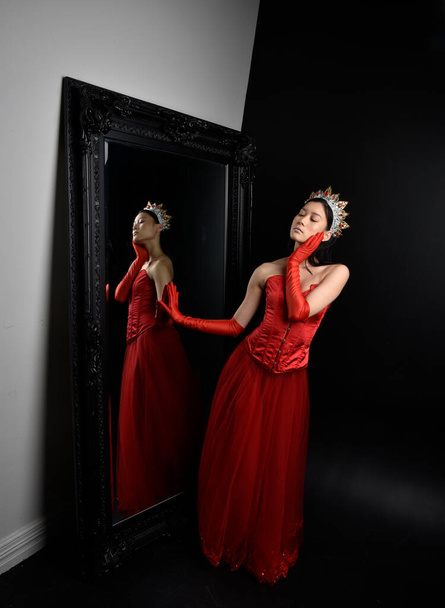 Ganztägiges Porträt einer schönen jungen Asiatin mit rotem Korsett, langen Opernhandschuhen und kunstvoller Kopfbedeckung. Anmutiges Posen vor einem Ganzkörperspiegel mit dunklem Studiohintergrund. - Foto, Bild